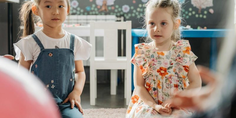 Two young girls sitting in a colorful kindergarten classroom, expressing innocence and curiosity.
